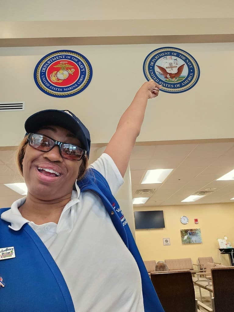 A Mu Alpha Rho founder in a VA volunteer vest raising her arm toward the Department of the Navy seal in a Veterans Affairs clinic