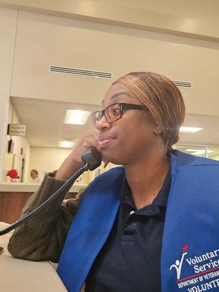 A Mu Alpha Rho founder in a VA Voluntary Service vest answering a clinic phone line at a Veterans Affairs clinic